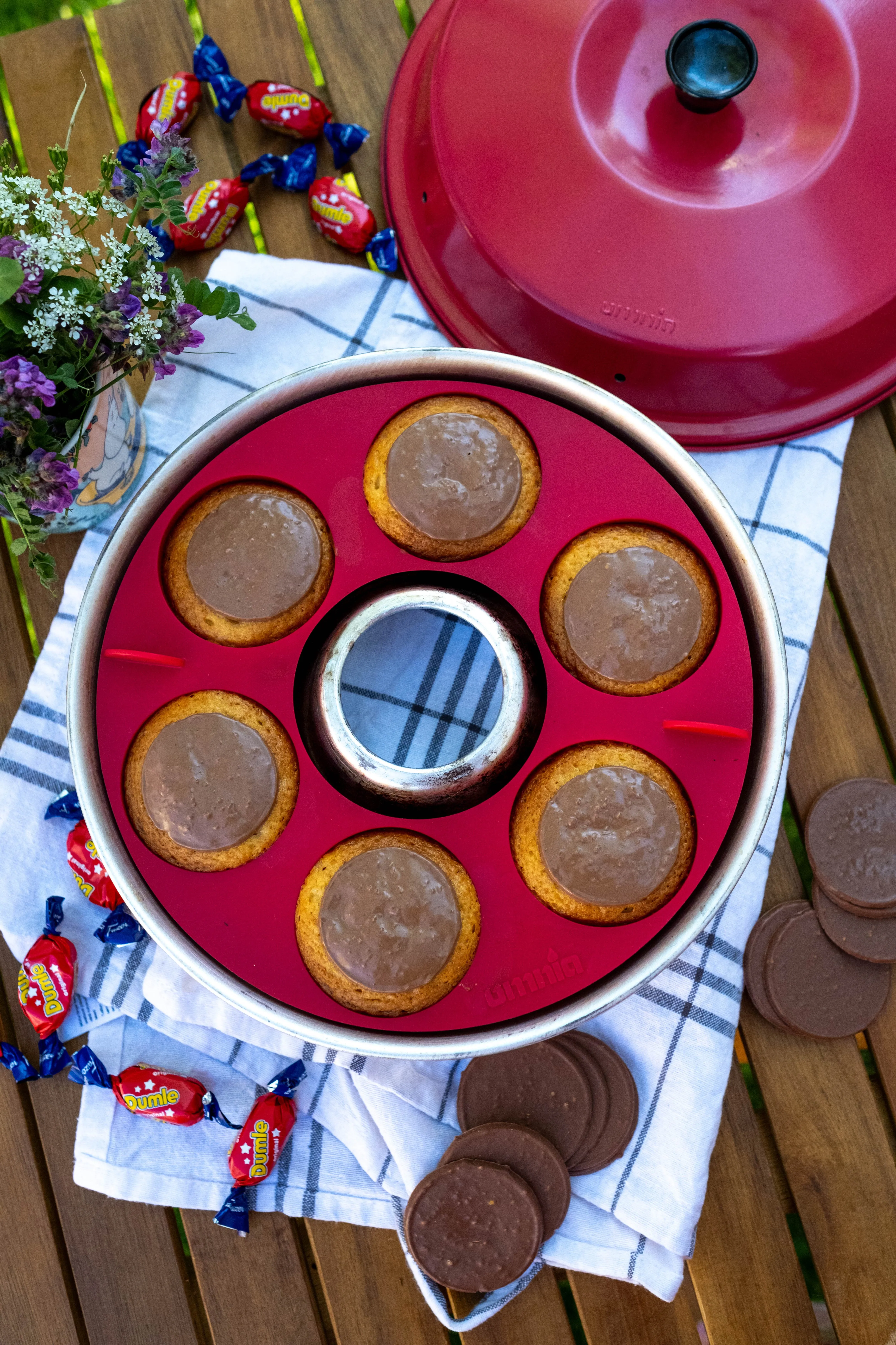 Roter Muffin-Tab aus Silikon mit mehreren goldbraunen, glasierten Muffins auf kariertem Tuch, umgeben von Blumen und Süßigkeiten