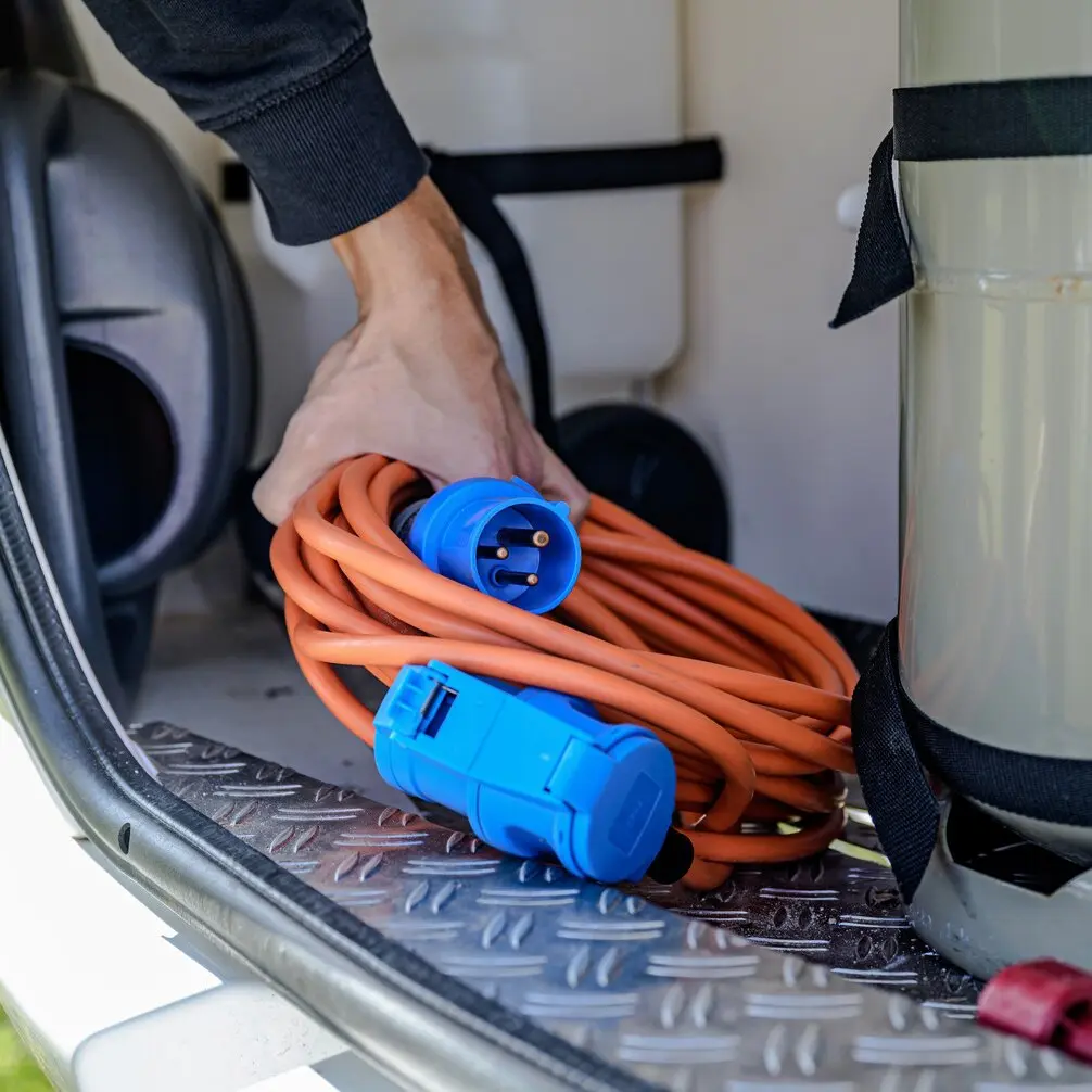 Orangenes Camping-Verlängerungskabel mit CEE-CEE/Schuko-Steckern, 25 m, aufgerollt; blaue Stecker, Hand greift danach, Diamantmuster-Boden im Fahrzeug