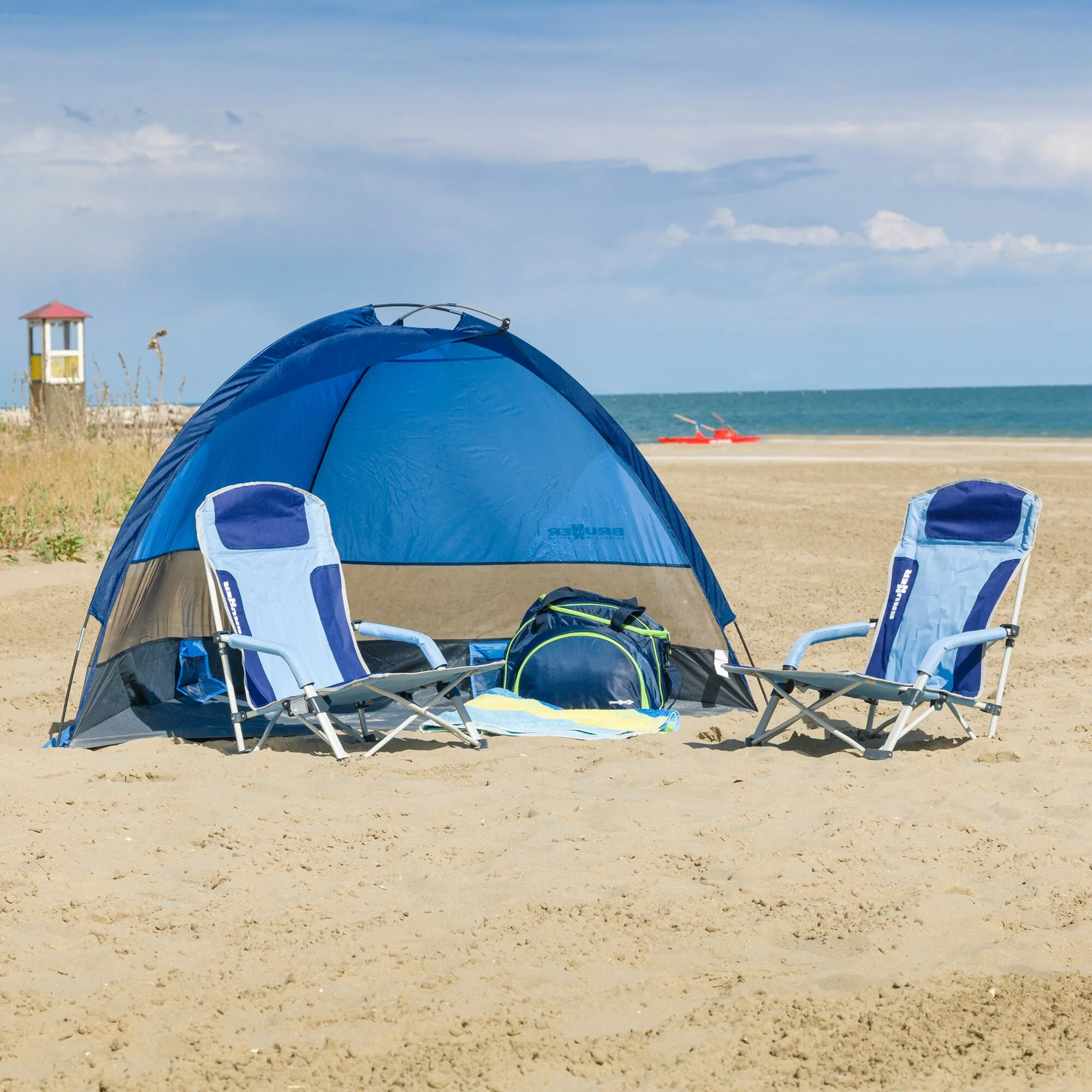 Zwei blau-weiße faltbare Strandstühle Bula stehen vor einem blauen Strandzelt an einem Sandstrand mit Meerblick