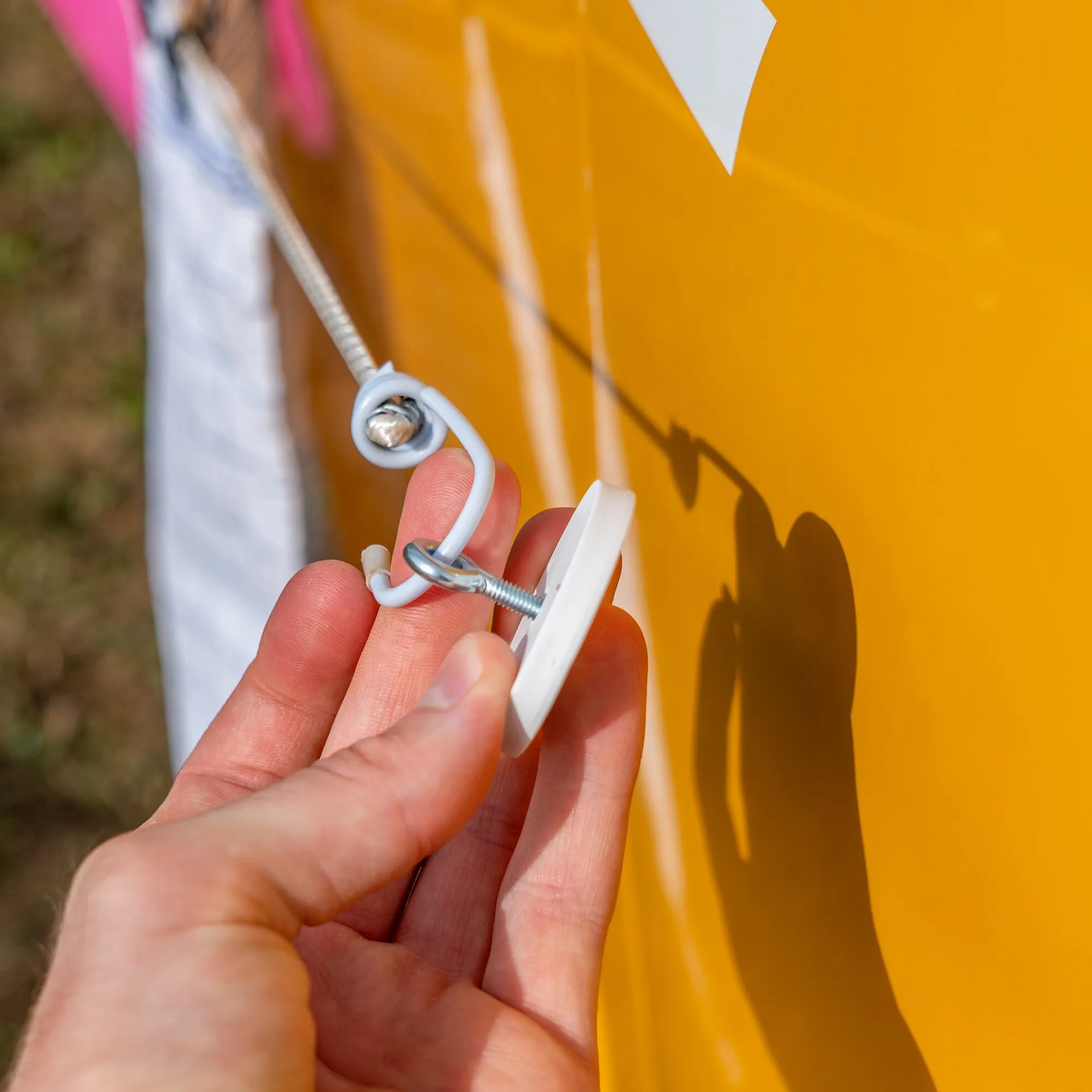 Hand hält ein weißes Magnet-Set mit Schraube und Metallöse vor einer orangefarbenen Fläche gras