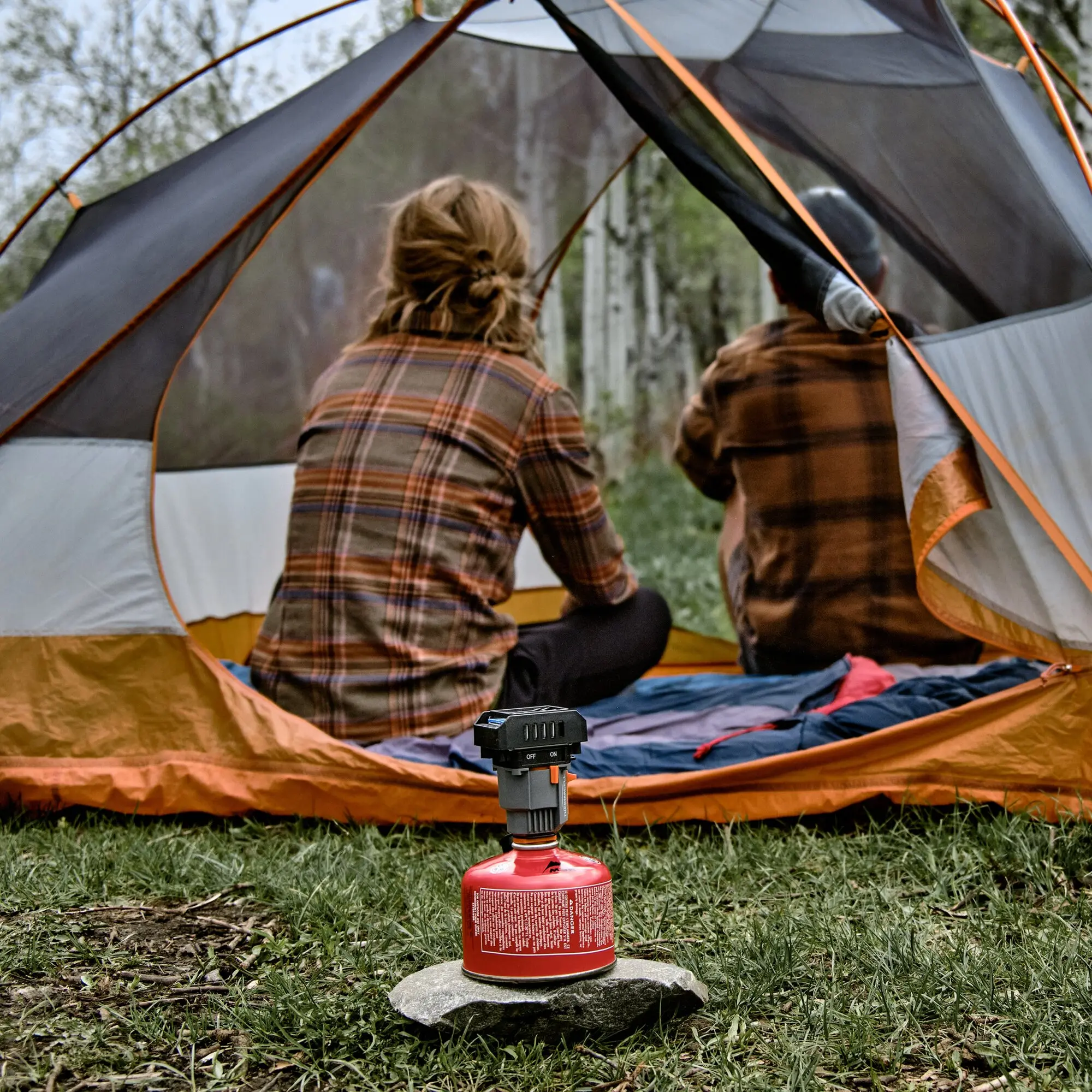 Zwei Personen in karierten Hemden sitzen im geöffneten Campingzelt; davor steht eine rote Propangasflasche mit Regler und On/Off-Schalter