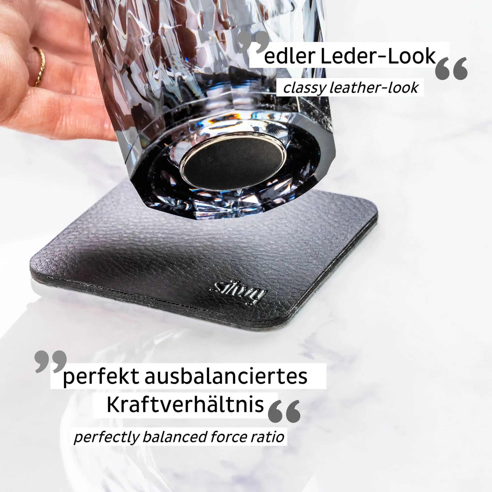 Graues Kunststoff-Whiskyglas mit facettierter Oberfläche, schwarzem Magnetboden und Leder-Look-Untersetzer in einer Hand