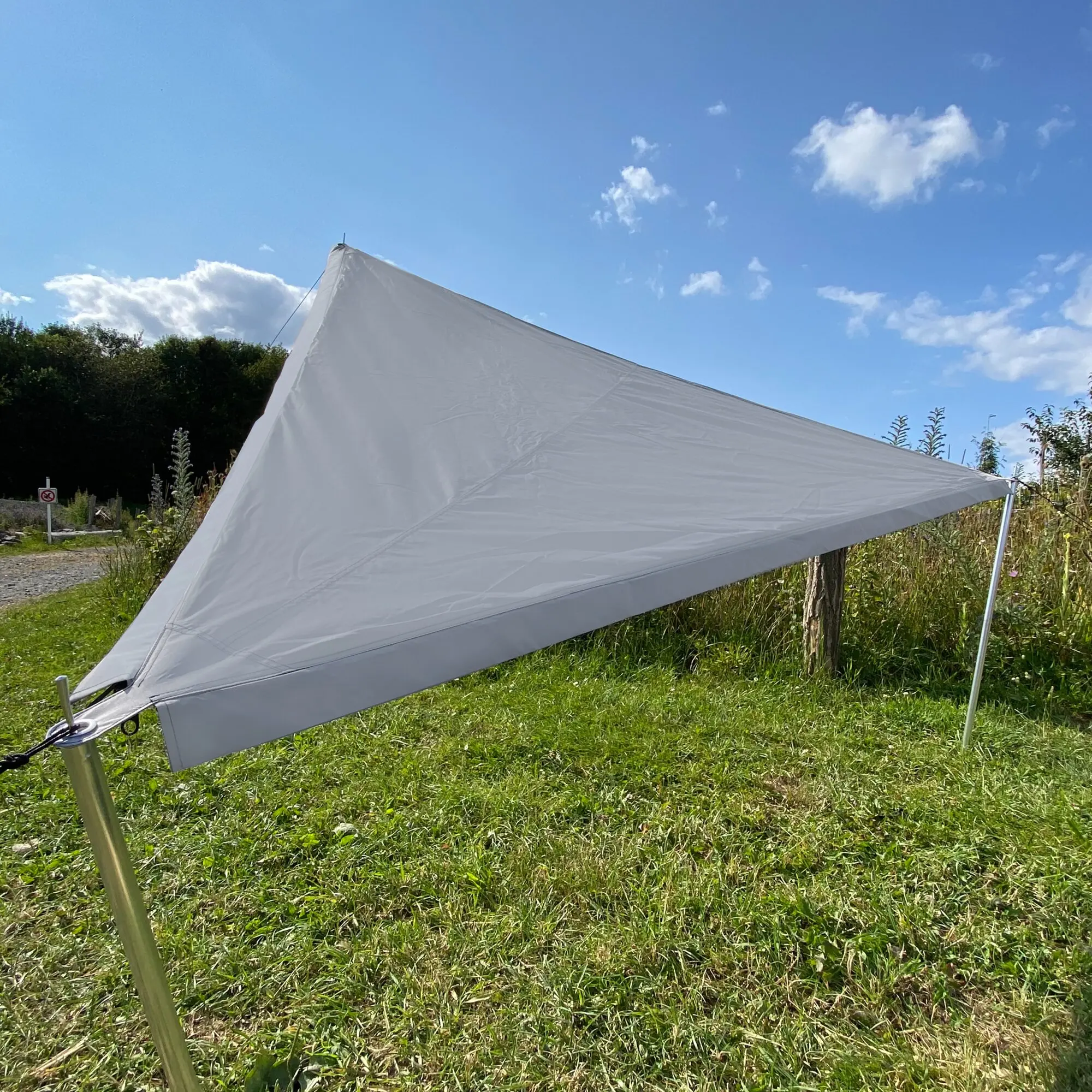 Weißes Dreiecks-Sonnensegel aus Canvas, gespannt zwischen Pfosten, Seile und Stangen, Gras und Gebüsch im Vordergrund, blauer Himmel