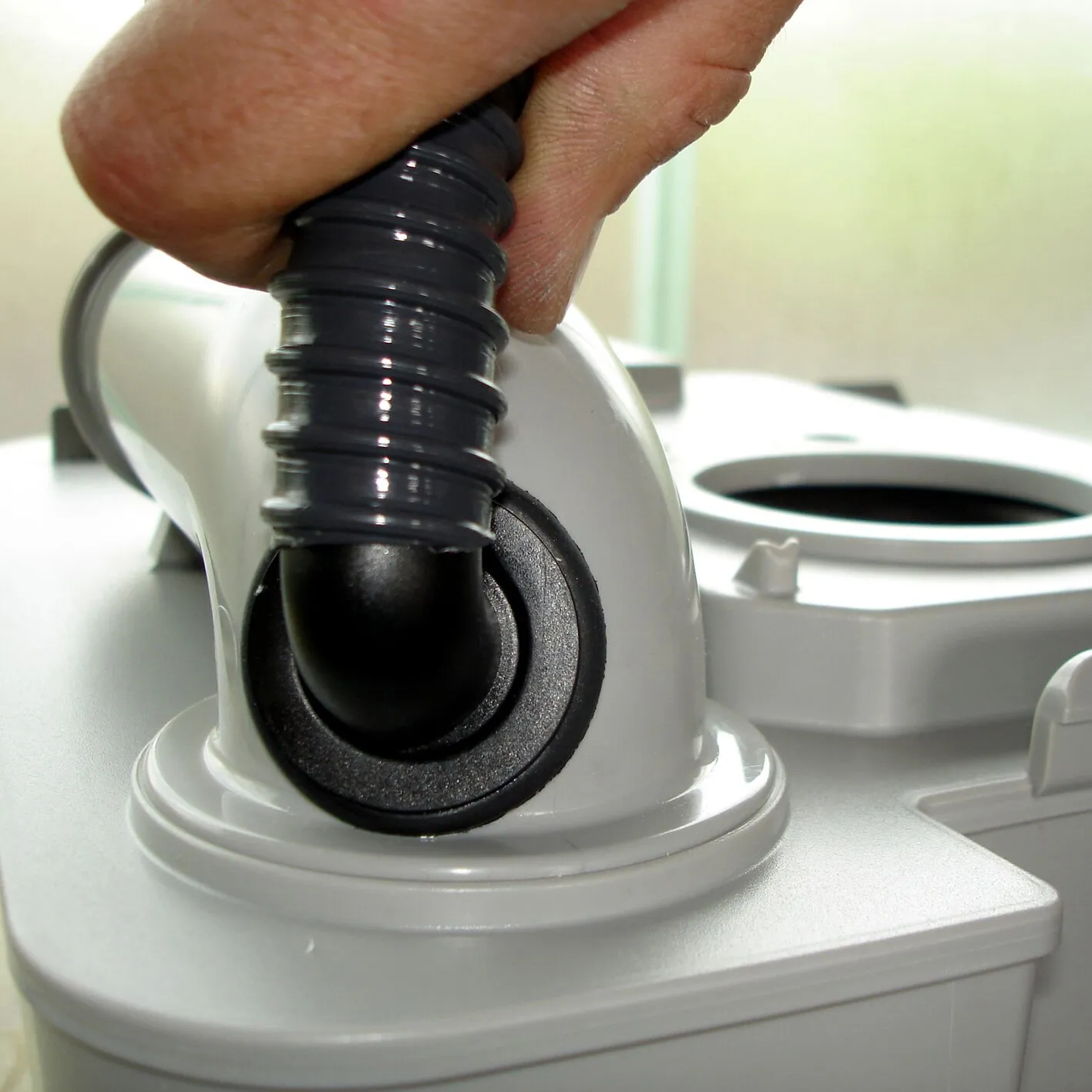 Detailaufnahme einer weißen Porta-Potti-Toilette mit schwarzem Entlüftungsschlauch, der von Hand am Abluftanschluss befestigt wird