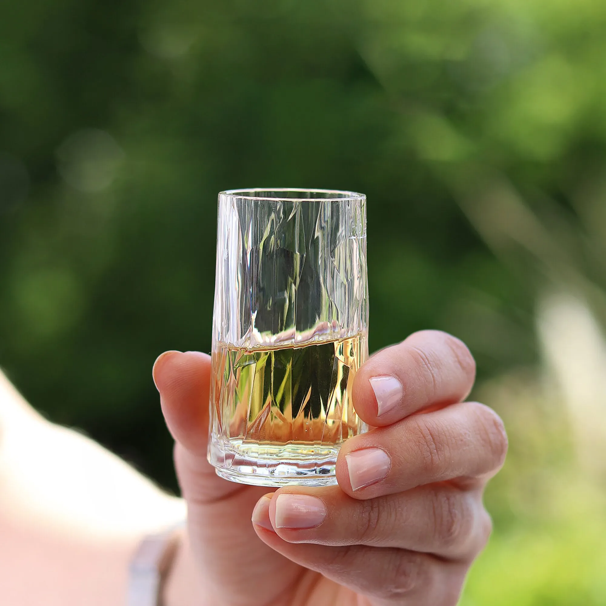Transparentes, zylindrisches Magnet-Schnapsglas aus Kunststoff mit goldenem Getränk, von einer Hand vor grünem Hintergrund gehalten