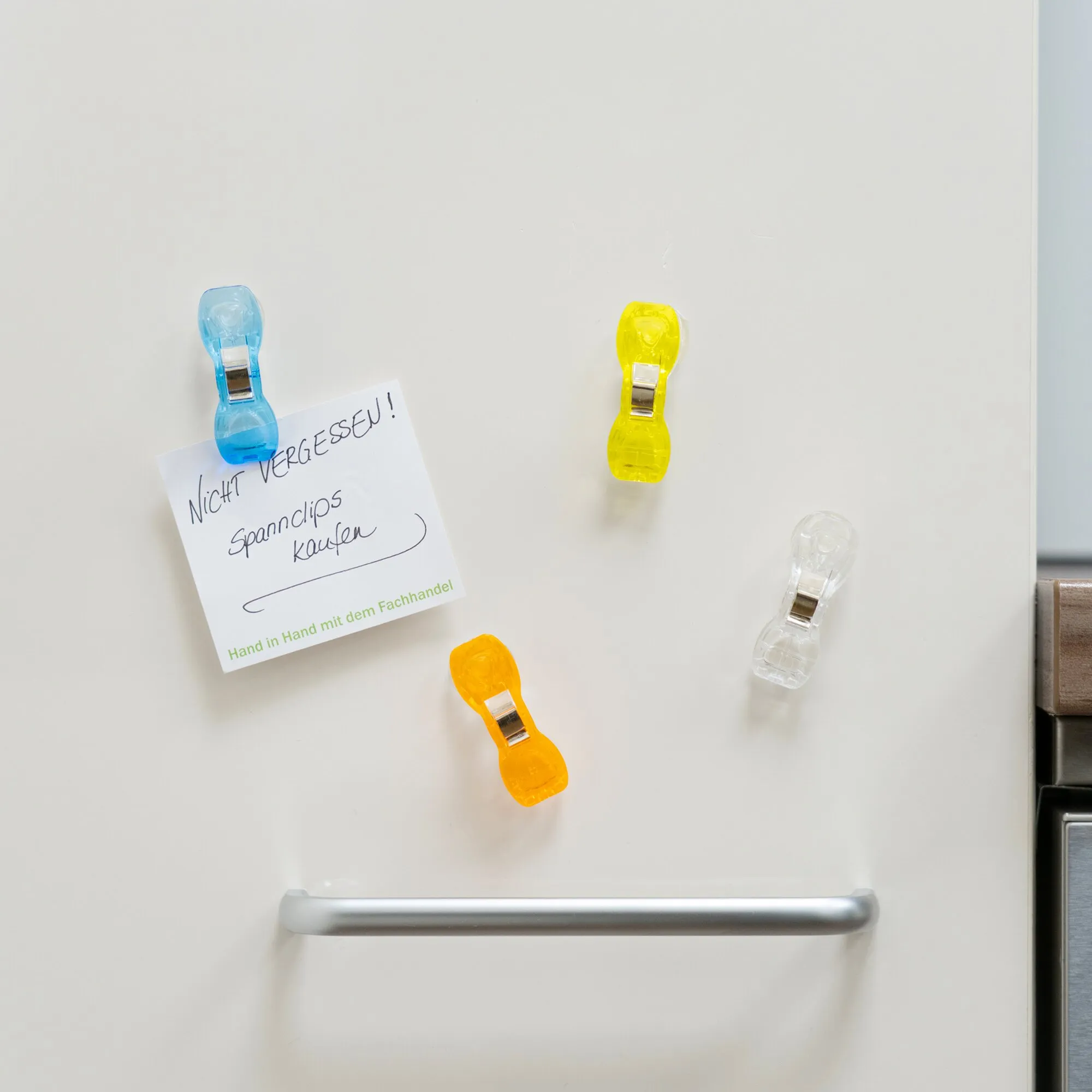 Mehrere bunte, transparente Spannclips mit Saugnäpfen kleben an einer glatten weißen Kühlschranktür; blau und gelb vorne, orange unten