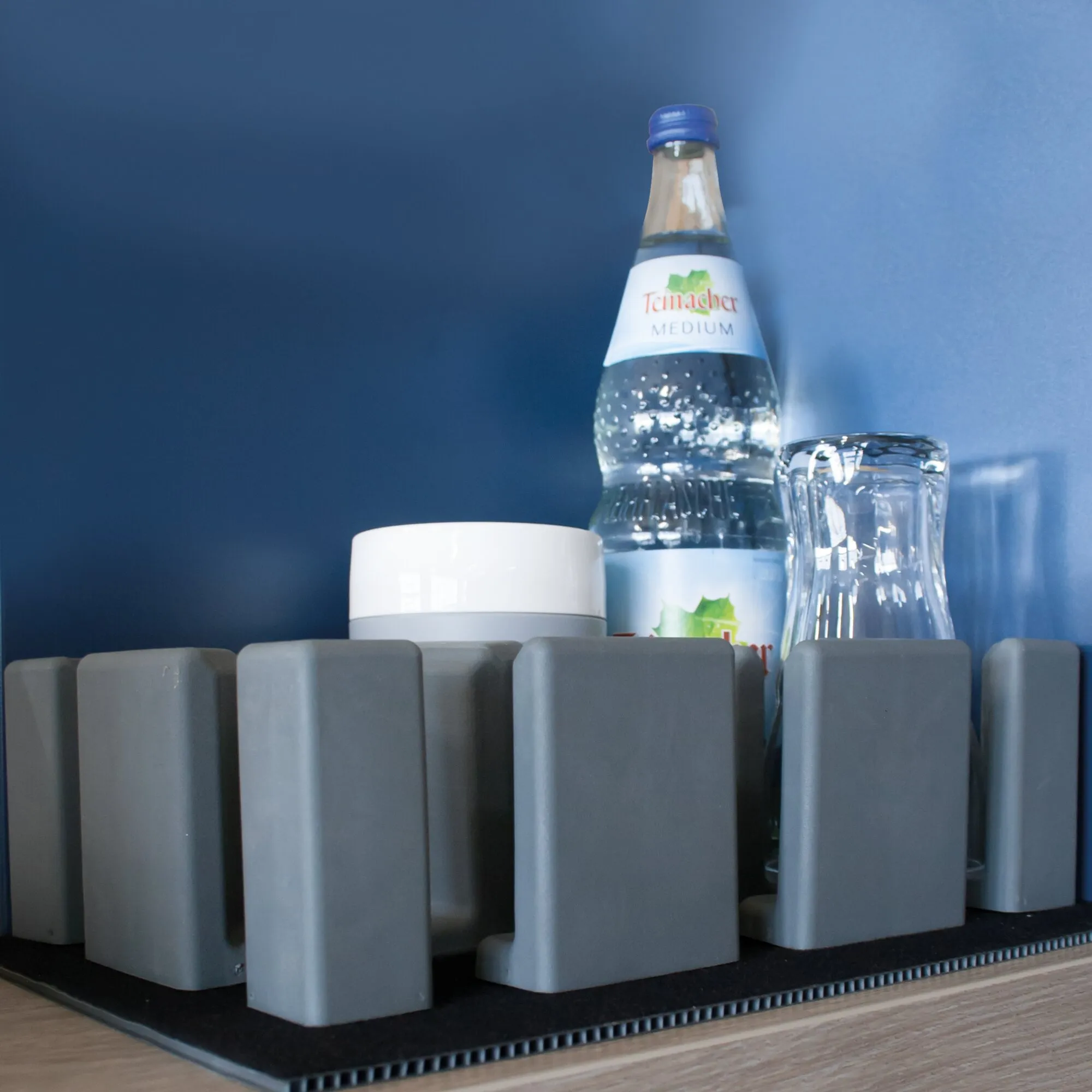 Graues Vario System Modul II mit modularen Halterungen für eine Mineralwasserflasche und ein Glas vor blauem Hintergrund