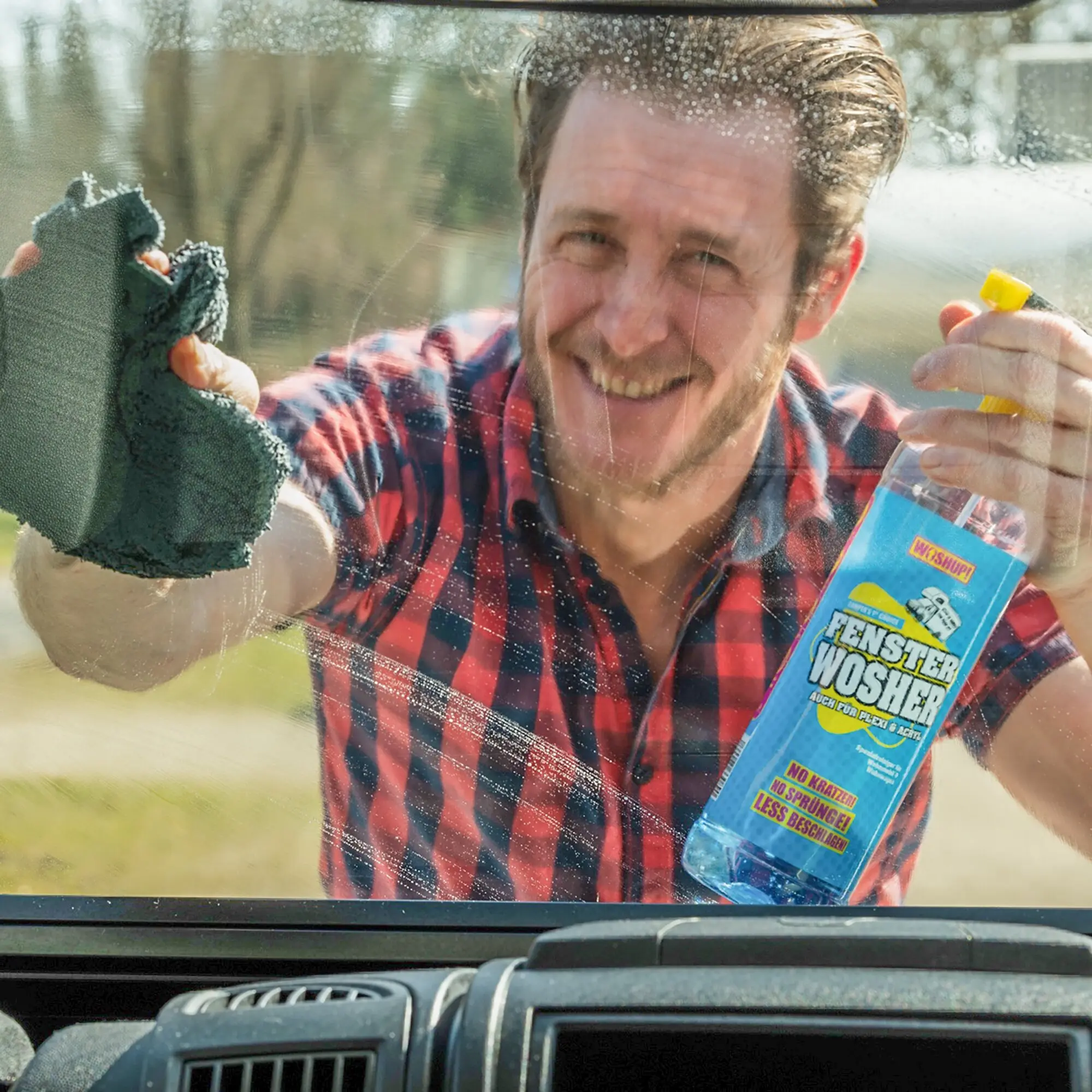 Fröhlicher Mann in rotem kariertem Hemd reinigt die Scheibe eines Autos, er hält gelbe Sprühflasche und grünen Schwamm, rechts blaue Fenster-Wosher-Verpackung