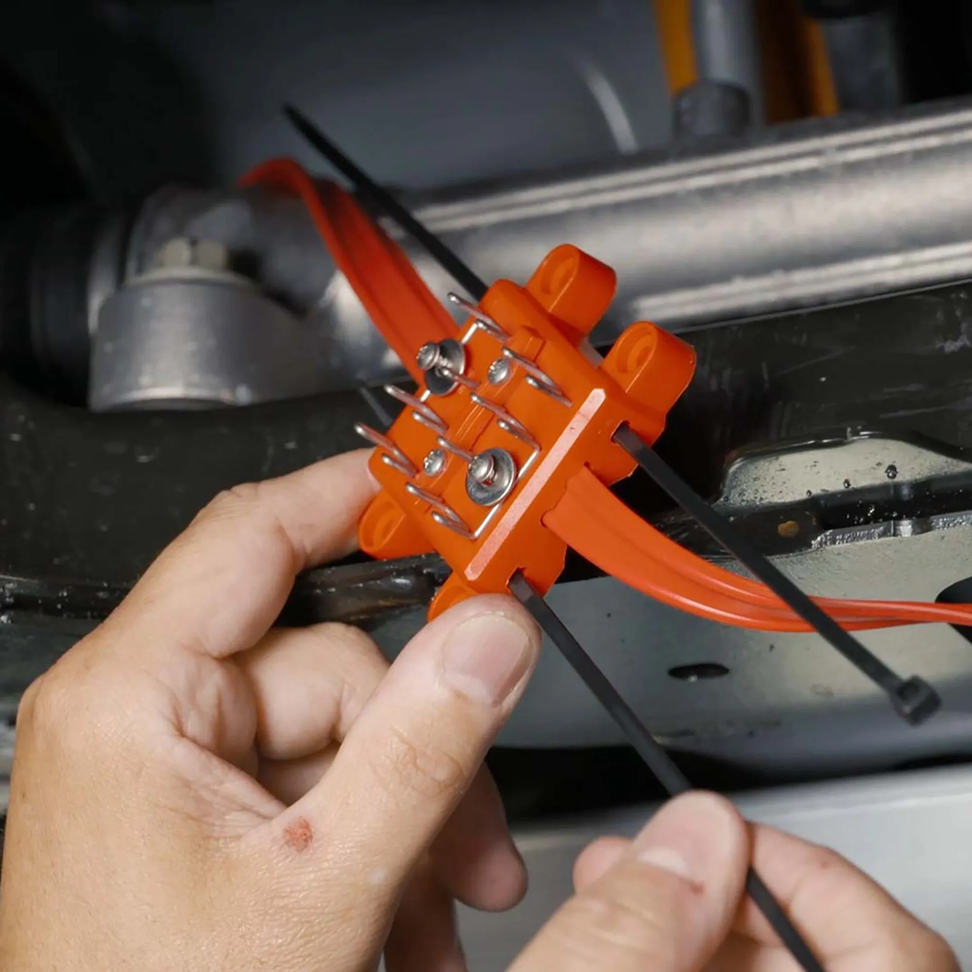 Orangefarbenes Marderabwehrgerät M9300 mit Metallkontakten und Kabeln, von einer Hand im Motorraum montiert