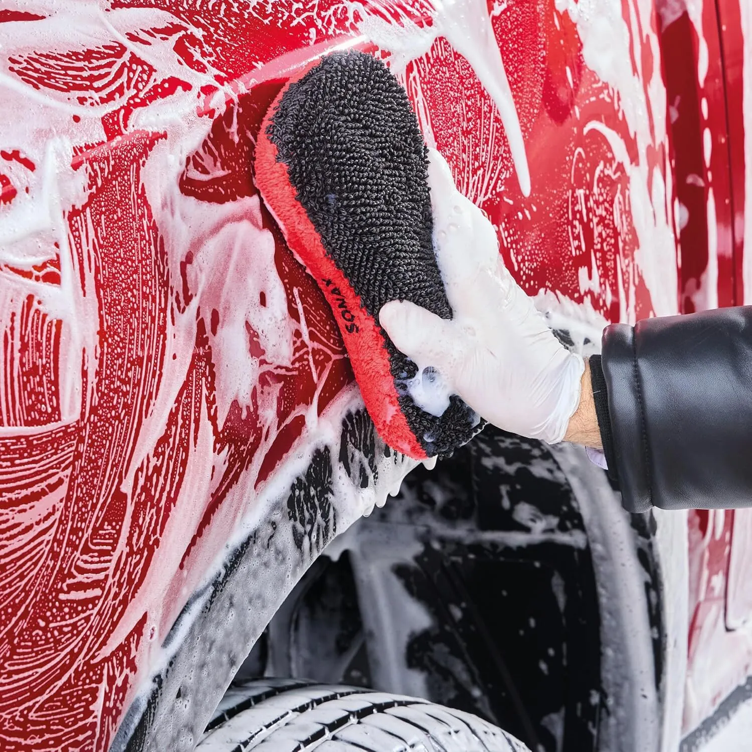 Schwarz-roter Microfaserschwamm wird von einer behandschuhten Hand zur Autowäsche auf ein rotes Fahrzeug aufgedrückt