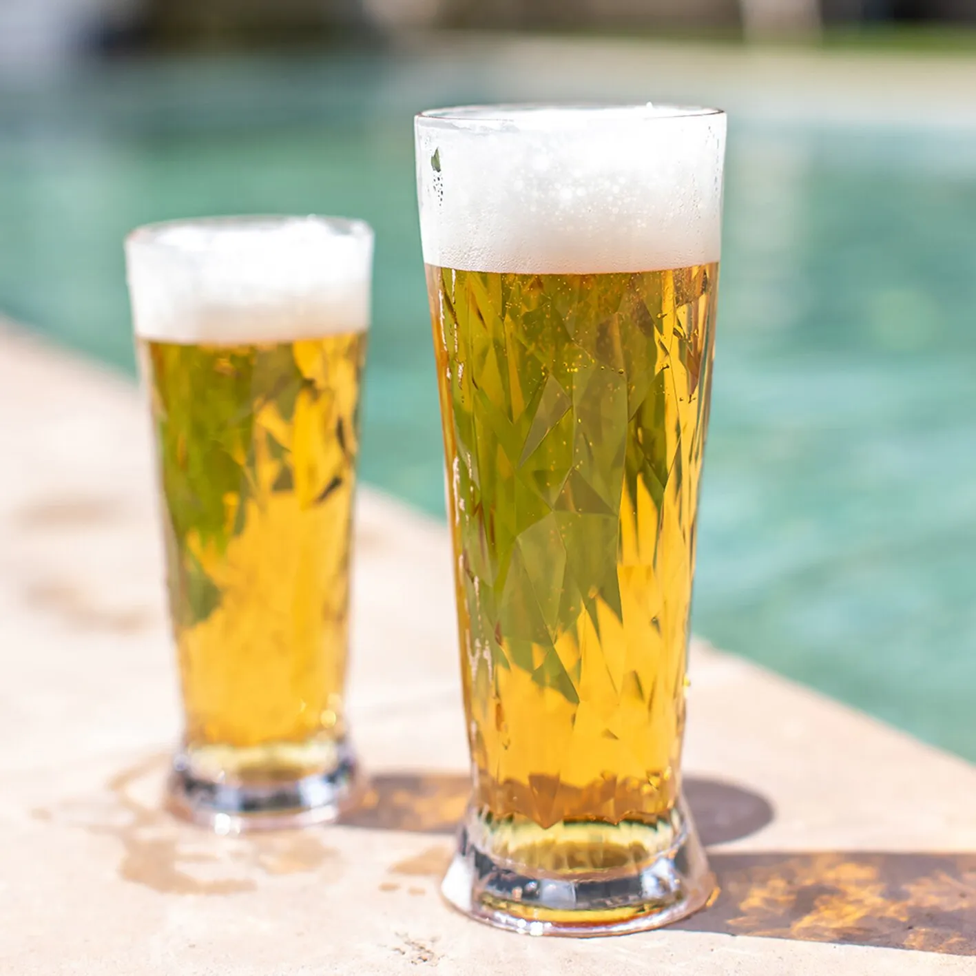 Zwei transparente Superglas CLUB Weißbiergläser mit goldenem Weißbier und weißem Schaum auf einem Tisch am Pool