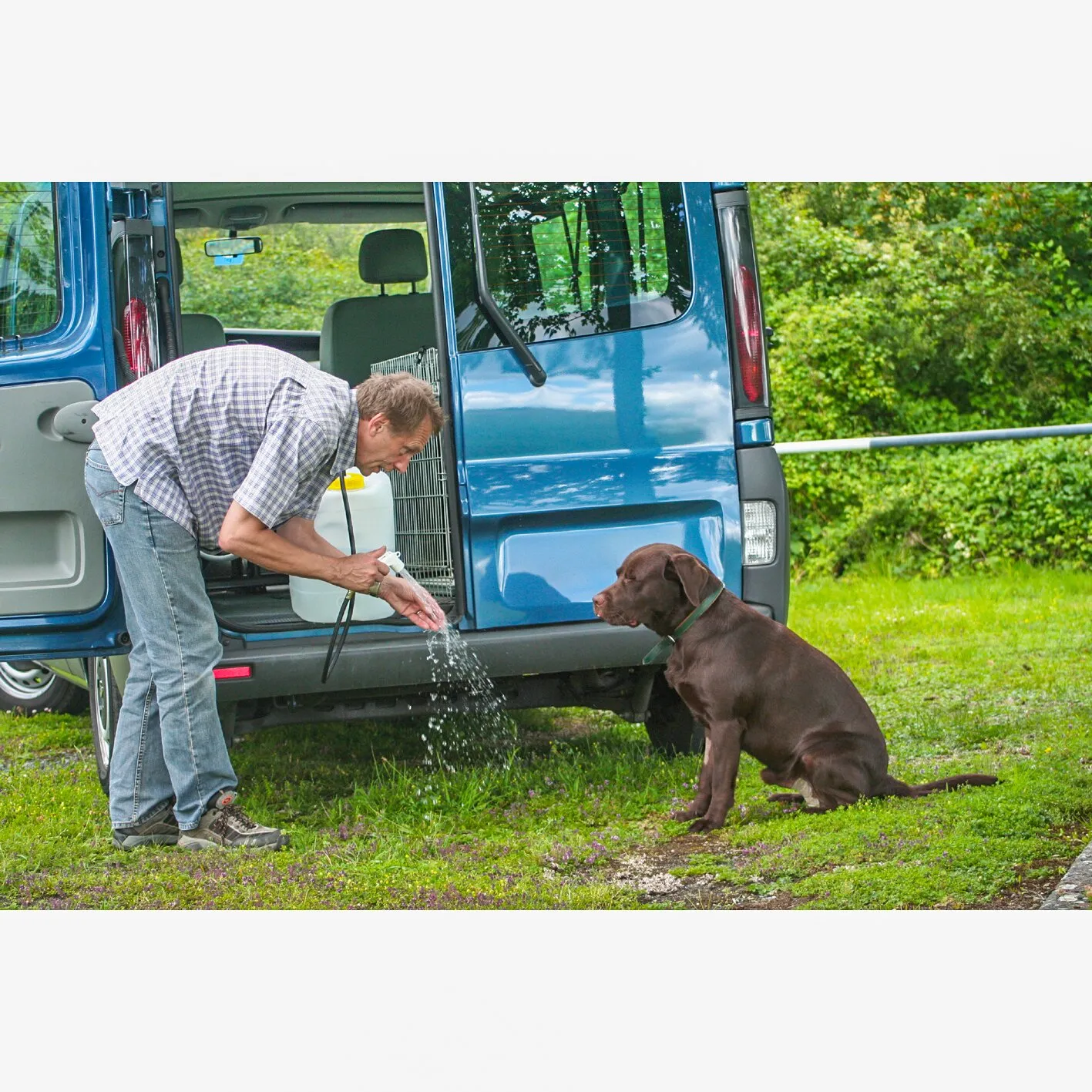Mann nutzt eine Kfz-Mobildusche am geöffneten blauen Fahrzeug, um einem sitzenden braunen Hund Wasser über die Hände zu spülen