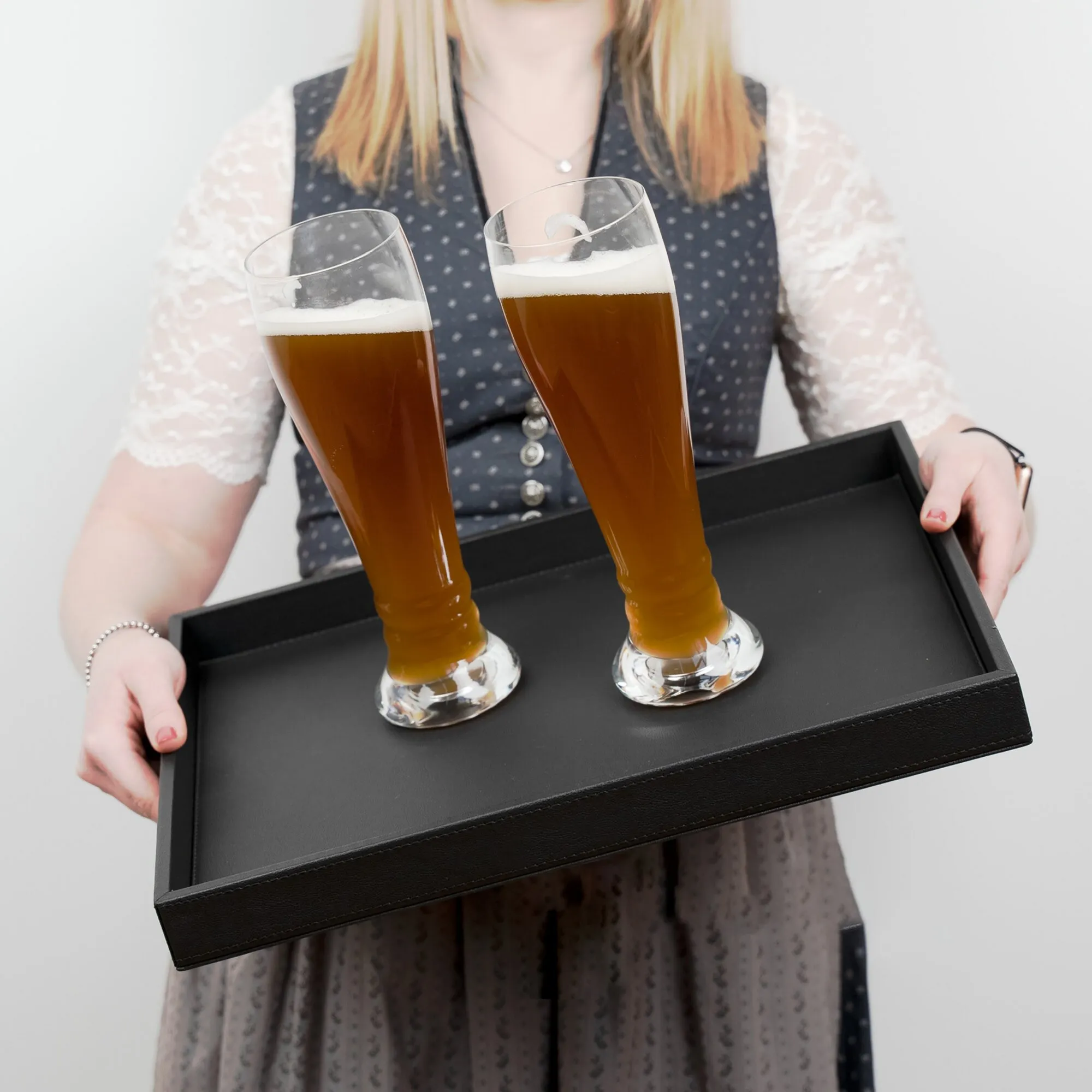 Zwei bauchige Weißbiergläser aus klarem Kristallglas mit dunklem Bier und Schaum auf einem schwarzen Tablett getragen
