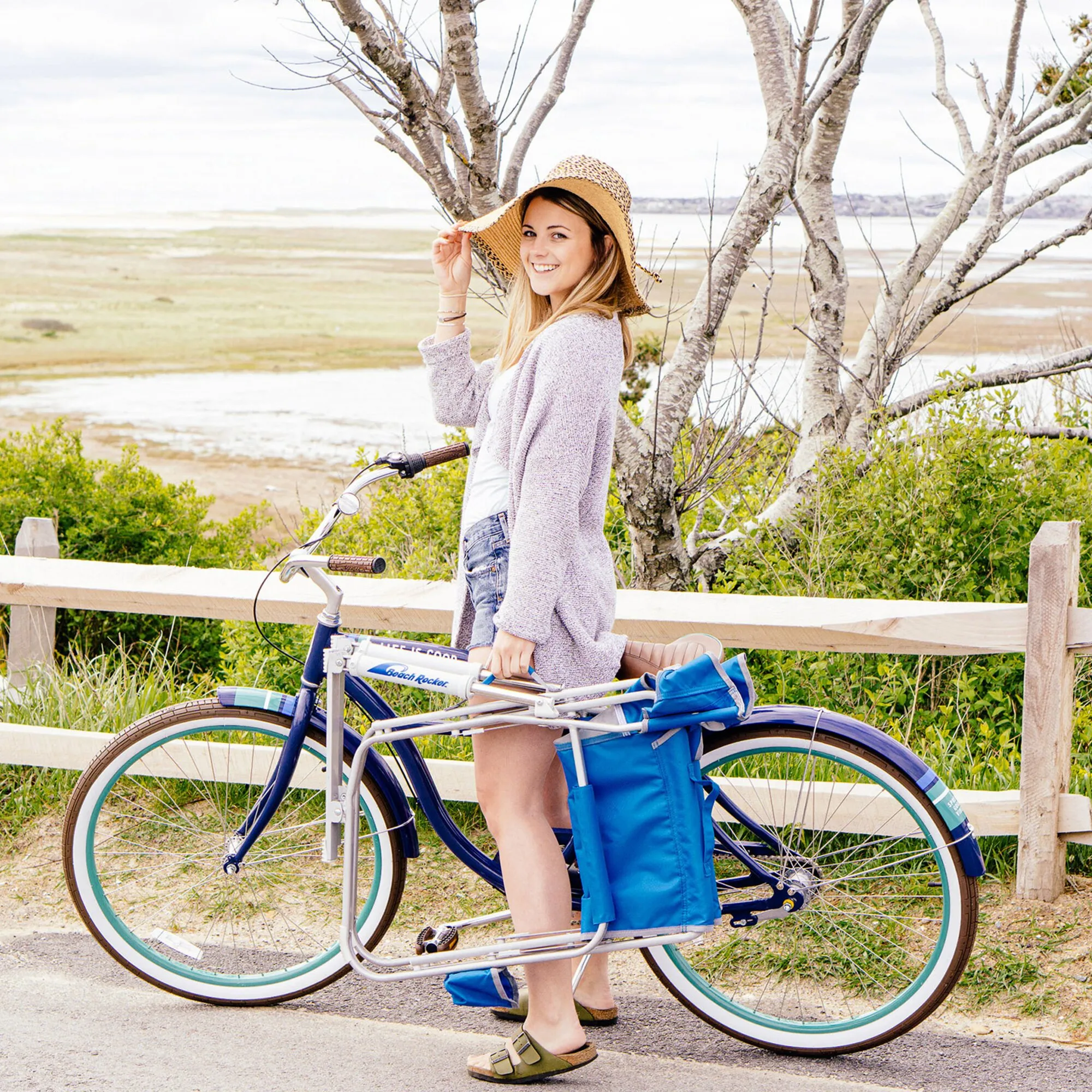 Frau schiebt ein Fahrrad am Strand, an dessen Gepäckträger der blaue Camping-Schaukelstuhl Beach Rocker™ befestigt ist