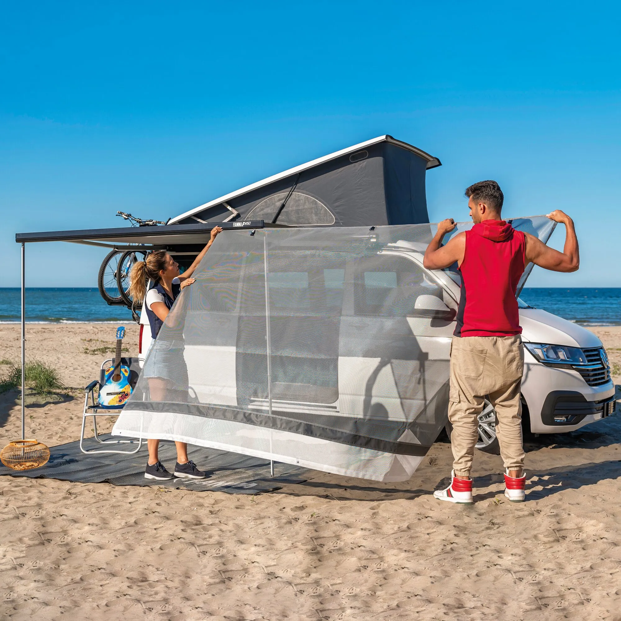 Vorderwand Sun View XL 375 aus grauem Mesh am Campingbus, während zwei Personen sie am Strand aufbauen